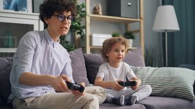 Happy family mother and son are playing video games in apartment having fun together, boy is jumping expressing positive emotions. Modern children and devices concept.