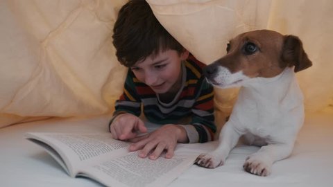 Distance learning. Online education. School. Happy boy reading emotionally book in pajamas dog Jack Russell lying bed under blanket. Child looks dog, reads book, running finger across. Pet. Quarantine