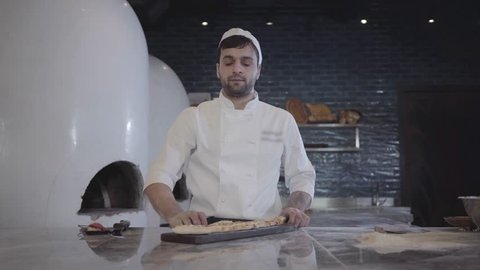 Portrait of chef in white uniform demonstrating freshly baked fatayer lying on the wooden board cutted on pieces in modern restaurant kitchen and looking in camera. Big oven is in the backgound