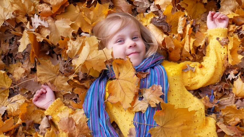 Smiling happy little girl lying and tossing or throwing fallen leaf in autumn in park