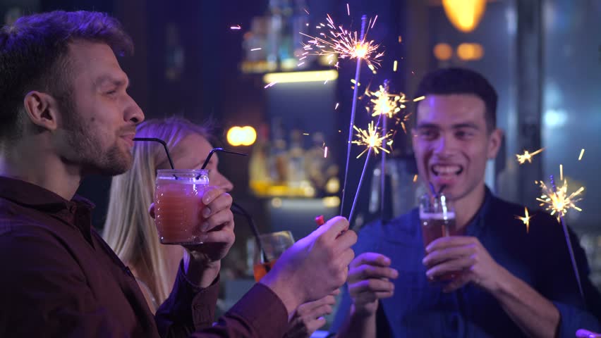 Happy group of diverse friends enjoying delicious cocktails, lighting sparkles at Birhday party. Cheerful nightclub visitors celebrating, clincking glasses, toasting at bar counter
