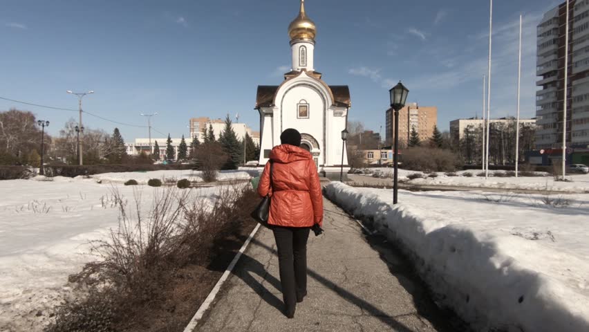 Young woman in a red jacket. Walking along the sidewalk in the city to the chapel. Warm spring day. Shooting from the back.