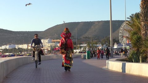 AGADIR, MOROCCO - MARCH, 2016: Rear view of water seller carrier dressed in traditional red costume walking away from camera on Agadir city seafront promenade