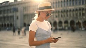 Caucasian young woman tourist in blue dress walking on square in Rome checking mail from friend on smartphone connected to 4G in roaming. Female blogger typing text of publication on website