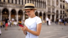 Happy pretty caucasian woman in hat and glasses enjoying journey during vacations walking with smartphone for networking and blogging via 4G roaming. Smiling female tourist using mobile phone 