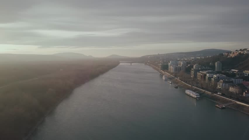 Aerial view travelling along river Danube skyline. Misty horizon to left & boat & hillside buildings to right frame. colourful sunrise landscape, Slovakia.