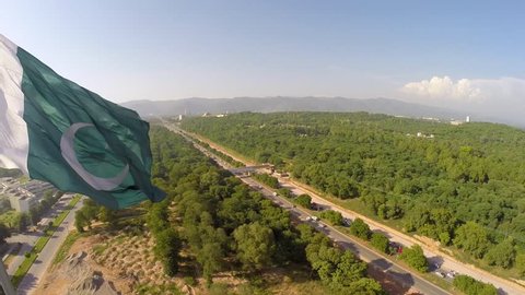 Islamabad, Pakistan - March, 2019: Beautiful Pan Right Aerial View of Weaving Flag of Pakistan with Green Trees and Margalla Hills at the Background 
