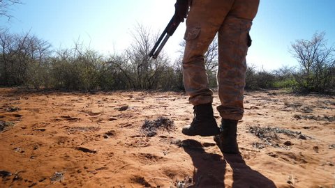 Low following shot with armed African ranger's legs in right of frame as he is identifying tracks in pursuit of poachers in the sandy bush
