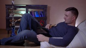 A young guy smokes a hookah, exhales smoke and talks on video by laptop. A young man in the smoke. A young guy on a pillow against the background of a bookcase and holds a phone in his hands.