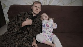 Cute small daughter with her funny father in bathrobe watching interesting film on tv with great emotions. Cheerful Caucasian father together with his cute daughter on sofa at home. Inside
