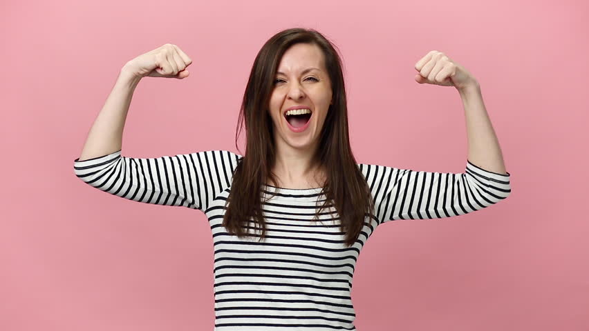 Smiling fun brunette young woman in striped shirt looking camera with charming smile, showing biceps on hand isolated over pastel pink background in studio. People sincere emotions, lifestyle concept. Royalty-Free Stock Footage #1026788855