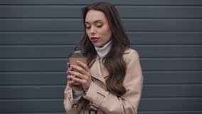 smiling elegant woman in beige trench coat drinking coffee from paper cup in cold windy weather