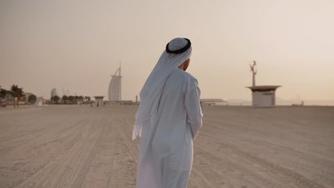 Caucasian man in white long clothes.Head is wearing keffiyeh.Walking throughsand,view is directed towards sea,which is visible from distance.In distance are visible buildings and silhouettes of people