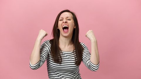 Overjoyed excited shocked brunette young woman in striped shirt looking camera say yes raising hands up in air isolated over pastel pink background in studio. People sincere emotions lifestyle concept