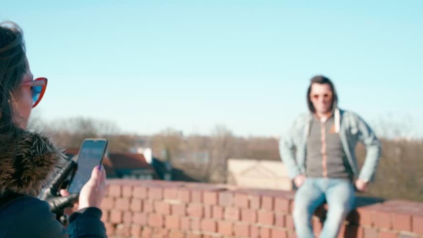 Over-the-shoulder shot of Woman Taking Picture of the Young Man Posing for Photo in the Old Town in Sunny Springtime Day
