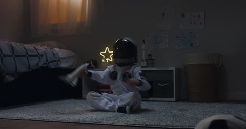 Cute little kid boy wearing astronaut suit playing with toy shuttle space ship rocket at home, bedroom interior evening shot. 4K UHD RAW FOOTAGE