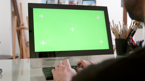 Man types continuously in front of a green screen. Artists workshop in the background. Designer working in his studio.
