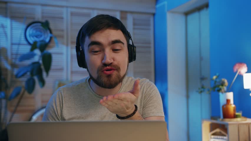 Young smiling man in headphones talking to girlfriend in video chat, using laptop. Nice bearded guy sending air kiss. Long distance relationships. Indoors. Device.? | Shutterstock HD Video #1026837650