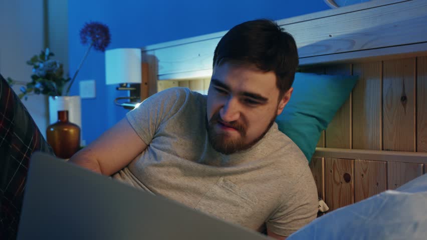 Good-looking smiling young man with beard looking at screen of laptop. Single guy chatting with girls in Internet at night. Dating site. Bedroom. Home. Indoors.?
