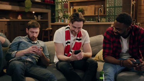 Multiracial group of friends looking at phone screen with smile while sitting on sofa by the tv. Close up diverse multiracial people sitting on couch using smartphones, addiction concept