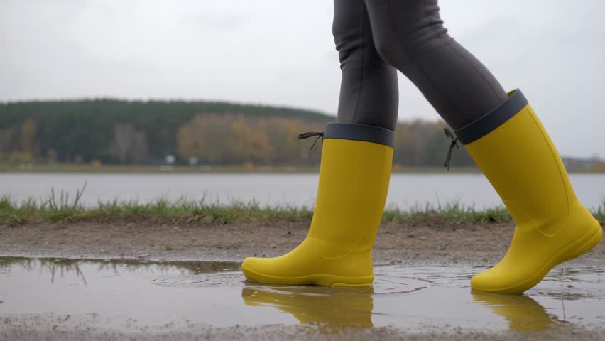 Legs of a woman in yellow boots walking through puddles and mud on a cloudy and rainy autumn day, unrecognizable person, slow motion