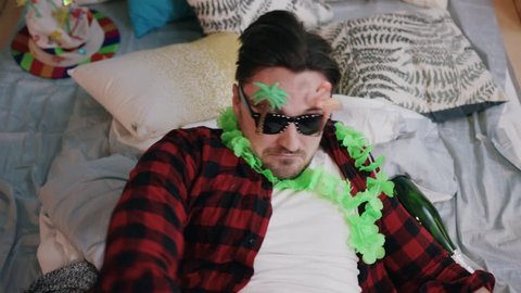 Young man in funny sunglasses and Hawaiian wreath laying in bed with pillows, sleeping. Guy awoke after party. Celebration concept. Hangover morning. Message, smartphone.