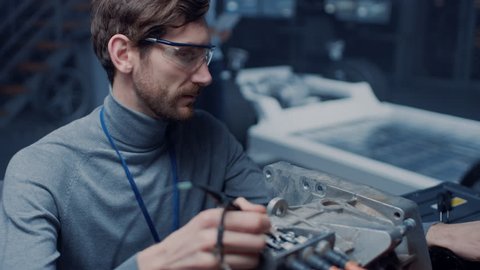 Professional Automotive Engineer in Glasses with a Computer and Inspection Tools is Testing an Used Electric Engine in a High Tech Laboratory with a Concept Car Chassis.