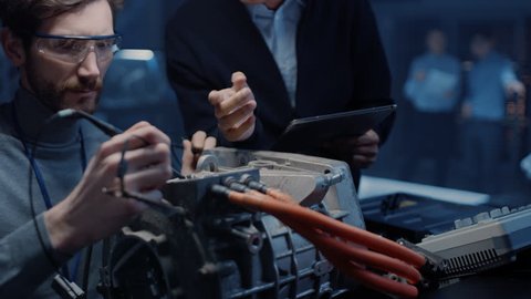 Two Professional Automotive Engineers with a Tablet Computer and Inspection Tools are Having a Conversation While Testing an Electric Engine in a High Tech Laboratory with a Concept Car Chassis.