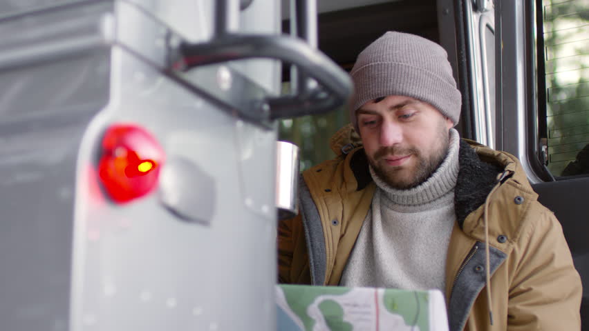 Panning of male tourist in warm clothes sitting in opened car with map in his hands, examining it and drinking tea from steel cup