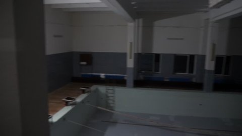 Empty indoor swimming pool drained with no water in an old school or prison building with lane ropes, a stair case and blue tiles with lights turning on