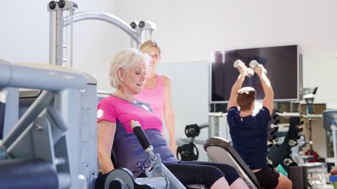 Senior Woman Exercising On Gym Equipment Being Encouraged By Personal Trainer 