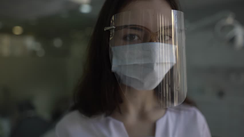 Close-up portrait of young female doctor who is wearing a dental shield, rounded around her face, from a frontal perspective to protect her glasses and eyes. Camera is slowly sliding. | Shutterstock HD Video #1026853052