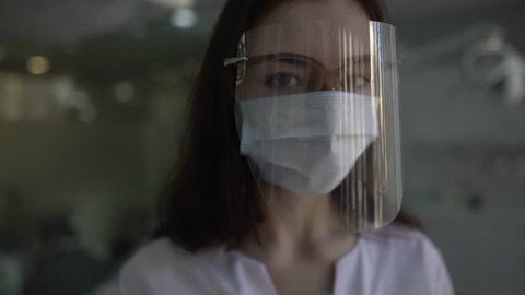 Close-up portrait of young female doctor who is wearing a dental shield, rounded around her face, from a frontal perspective to protect her glasses and eyes. Camera is slowly sliding.