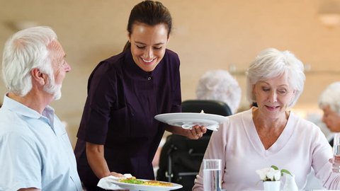Senior Couple Being Served With Meal By Carer In Dining Room Of Retirement Home