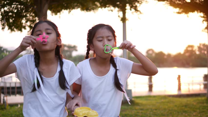 The old sister and young sister, who are wearing a white shirt, braid, cute white dresser, playing, blowing soap bubbles at the lawn on the riverside in the evening sunset.