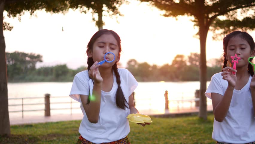 The old sister and young sister, who are wearing a white shirt, braid, cute white dresser, playing, blowing soap bubbles at the lawn on the riverside in the evening sunset.