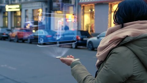 Unrecognizable woman standing on the street interacts HUD hologram with smartphone. Girl in warm clothes with a scarf uses technology of the future mobile screen on background of night city