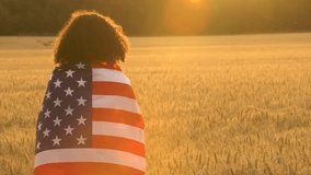 4K video clip of mixed race African American girl teenager female young woman wrapped in an American US Stars and Stripes flag in a wheat field at sunset or sunrise