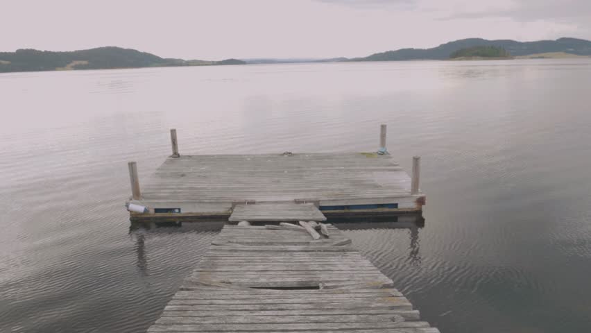 Man wearing a yellow rain coat carefully walking down onto a old board walk jetty with cracked wood overlooking a large flat calm lake and mountain ranges in the distance in Norway