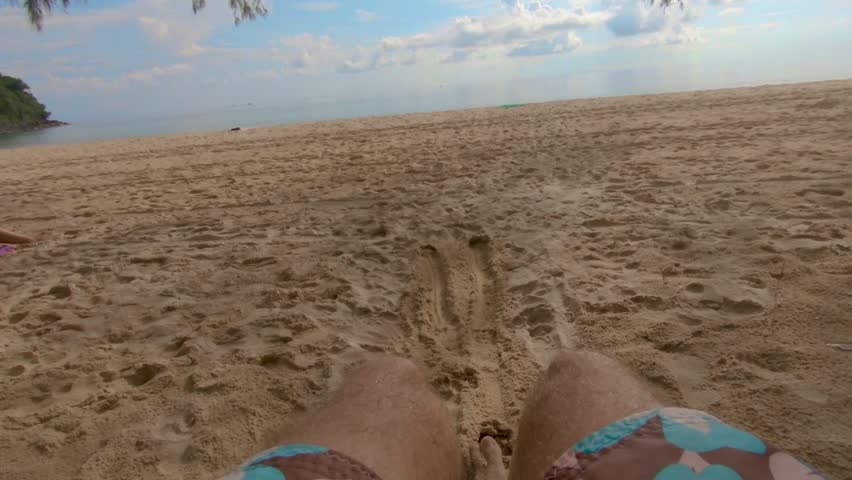 FPV slow motion of male feet swinging from a hammock in the beack with a colorful sky as background.