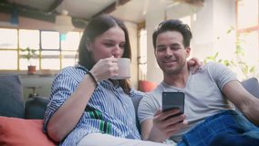 Young cute couple use new application on mobile device, enjoying to view online old family photos, connected to social media life, taking rest with cup of coffee in cosy living room
