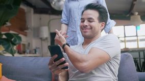 Happy Hispanic smiling couple family spend funny time together, watching online video on smart phone device, in cozy living room, girl hug her boyfriend, wearing watches, gray t-shirt and blue shirt
