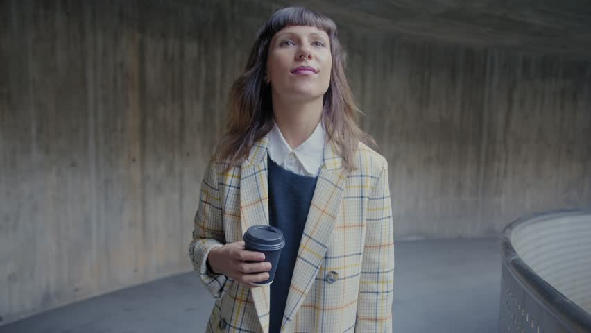 Young handsome pensive business woman walks around modern urban area, with take away cup, dressed in colorful suit, drinking hot coffee, looking at sunlight and thinking about future, concrete wall Royalty-Free Stock Footage #1026889070