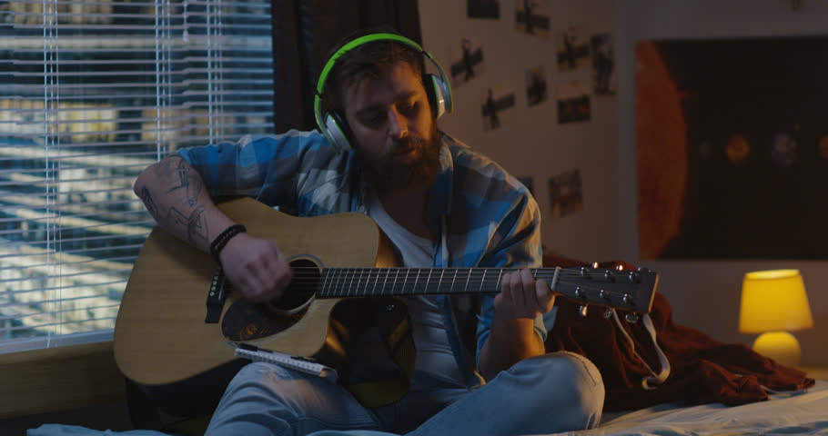 Medium shot of man sitting on bed and playing guitar