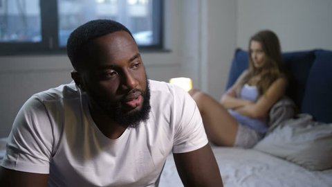 Close up shocked african american man discouraged by fail in bed due to problems in intimate sphere. Blurry on background sad woman frustratedly sitting on bed. Impotence is real relationship test