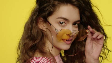 Playful woman in yellow glasses looking to camera. Portrait of fashion girl winking eye in studio. Close up of young woman in sunglasses winking to camera on yellow background. Fashion model wink eye