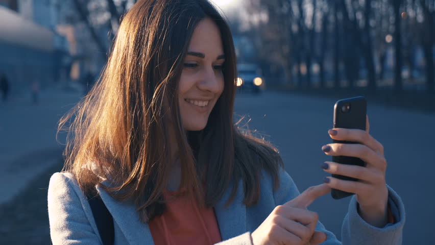 Attractive young woman walking at the sunny city streets , joyful girl using cellphone, sunset background.