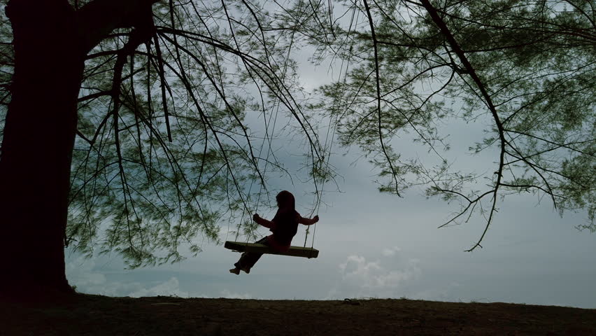 Silhouette of a Muslim girl plays a swing  under a pine tree on a beach - static shots, then pan upwards.
