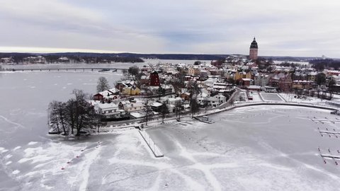 Drone footage flying towards a small town with a beautiful windmill the great old cathedral of Strängnäs. A frozen lake and a harbor is underneath. Filmed in realtime at 4k.