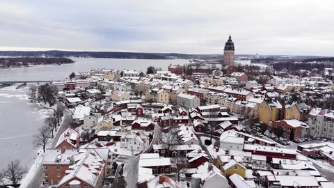 Drone footage flying over a small town towards the great old cathedral of Strängnäs. Colorful houses underneath and a frozen lake with a bridge in the background. Filmed in realtime at 4k.
